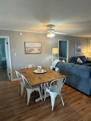 A dining area with a wooden table and white chairs, a ceiling fan, and a blue sofa in the background. - Oceanside - Quick 5 min walk to the beach - Great porch (Salvo)