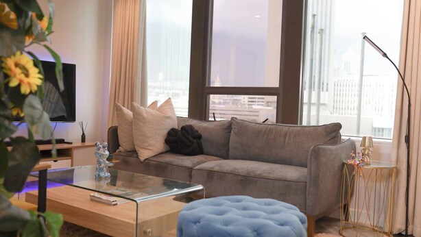 Ein modernes Wohnzimmer mit einem grauen Sofa, einem Glastisch, einem blauen Hocker und einem Fernseher auf einem Schrank.