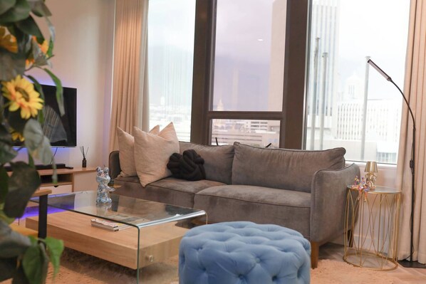 A modern living room with a grey sofa, a glass coffee table, a blue ottoman, and a TV on a stand.
