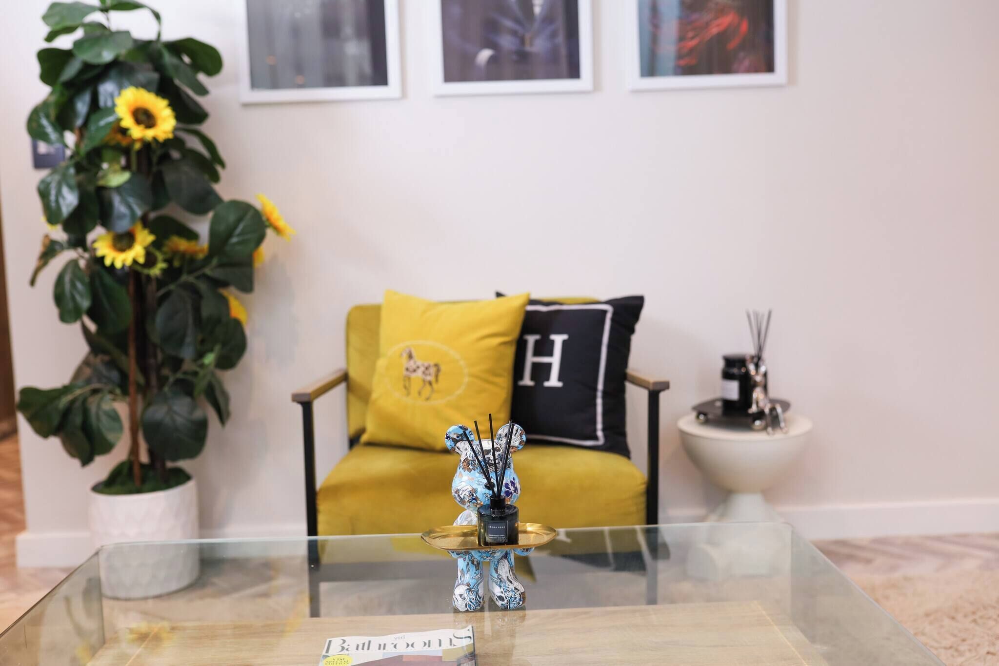 A living room with a glass coffee table, a sofa with yellow and black cushions, a potted plant, and a small table with a decorative item.