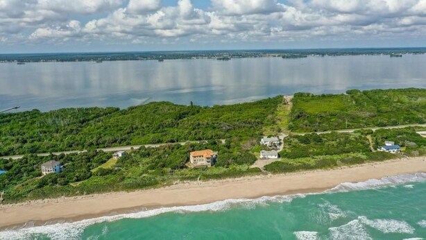 Vue aérienne d’une zone côtière comprenant une plage, une étendue d’eau importante et un paysage verdoyant dense parsemé de bâtiments.