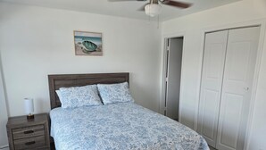 Une chambre à coucher avec un lit en bois, une table de chevet avec une lampe, un ventilateur de plafond et une porte sur le côté droit.