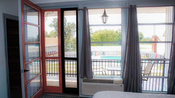 Una habitación de hotel con una cama, un ventanal corredizo que da a un balcón con piscina y mobiliario de exterior.