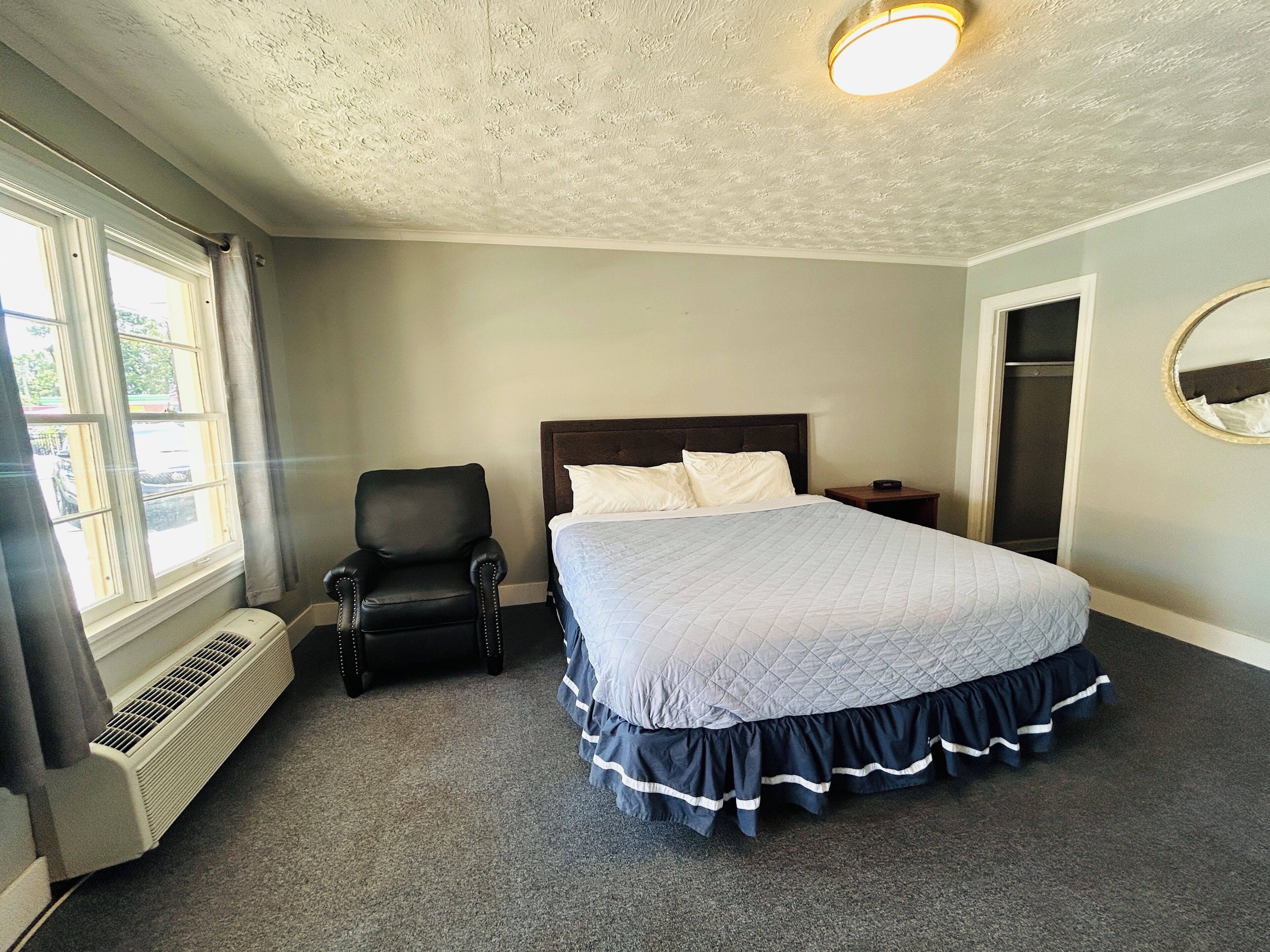 A bedroom with a bed, a chair, a nightstand, a mirror, and a window with curtains.