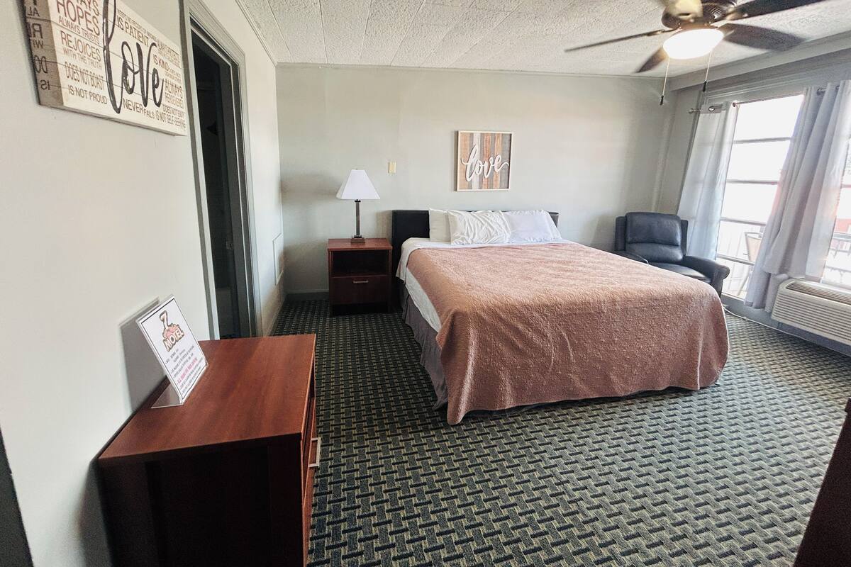 Une chambre d’hôtel avec un lit, une table de chevet, une lampe, une chaise et un ventilateur de plafond.