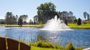 A lakeside view with a fountain, chairs, and a grassy area.