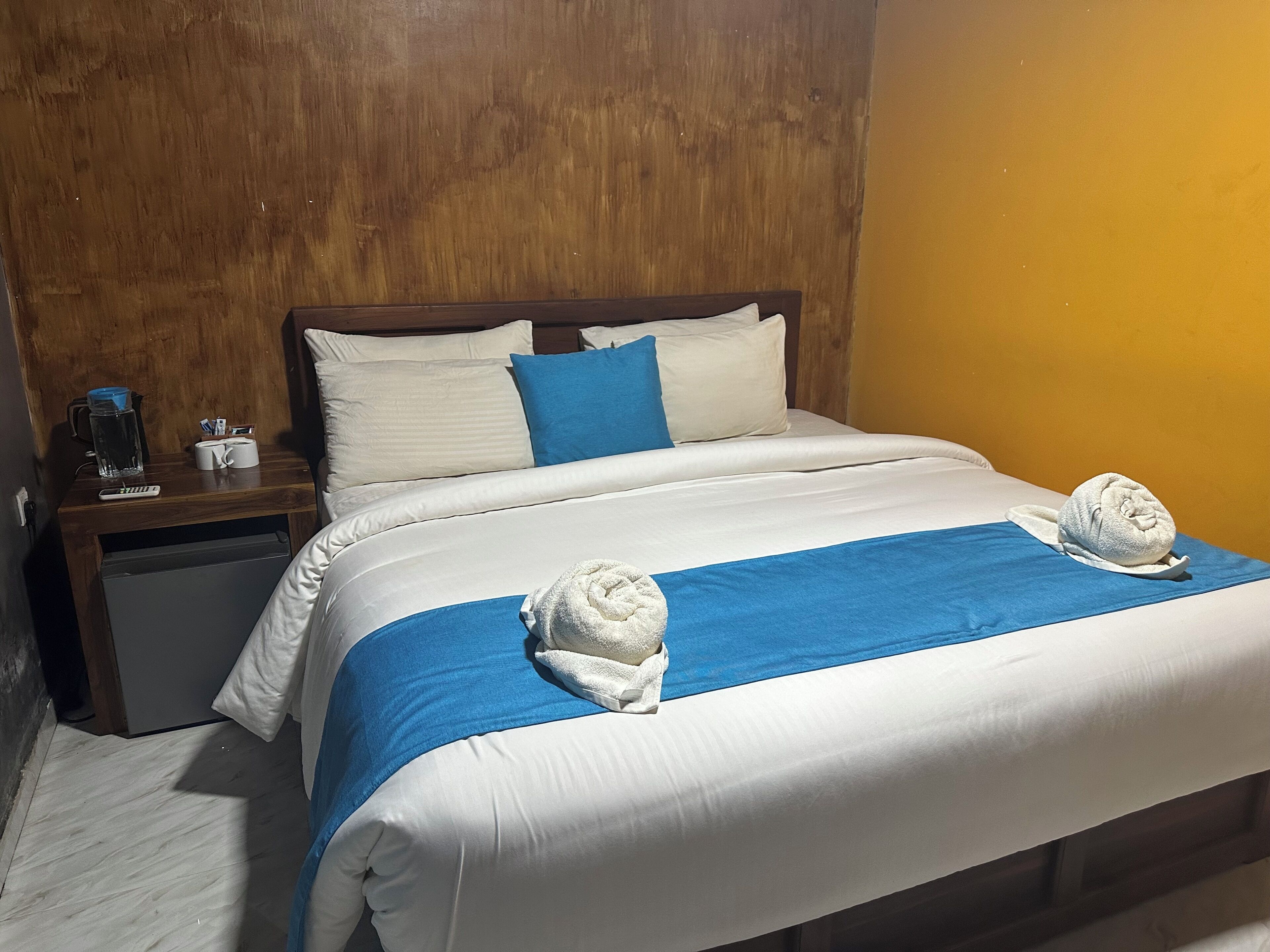 A hotel room with a bed, pillows, a nightstand, a water pitcher, and a towel.