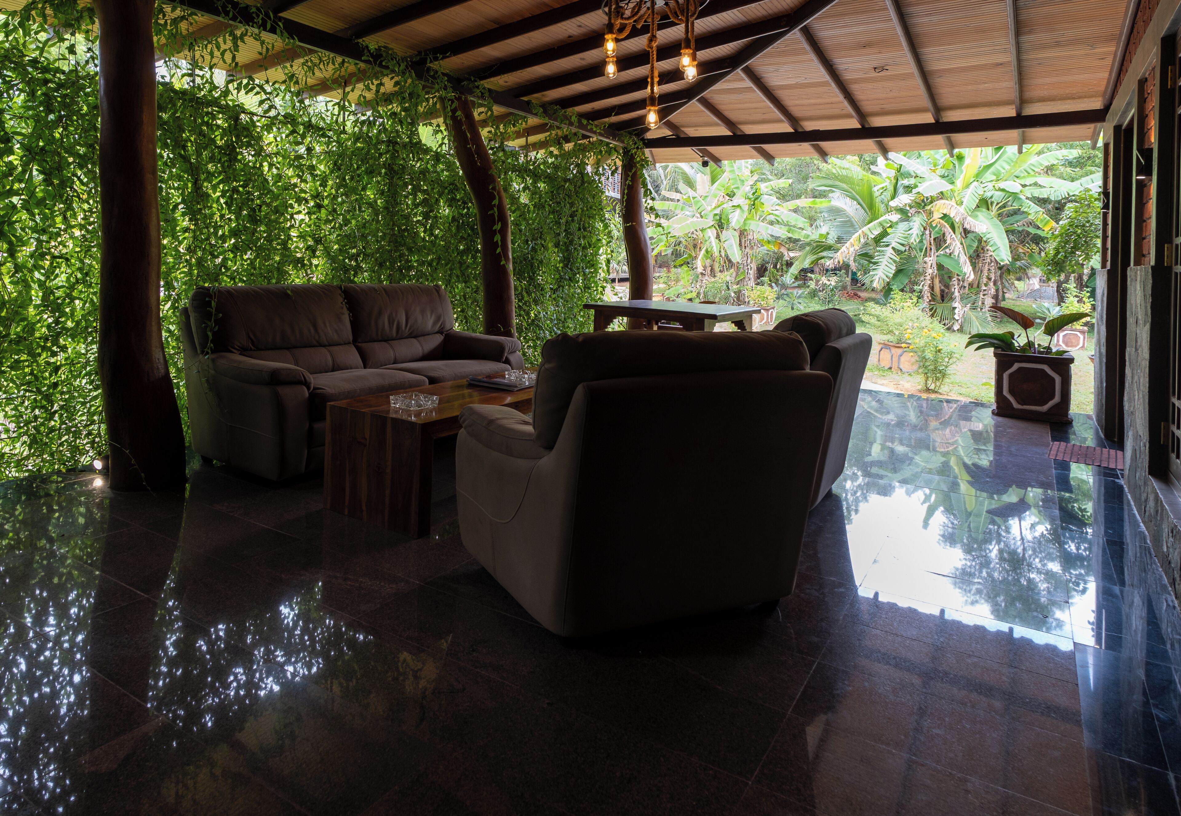 A covered outdoor seating area with a cushioned sofa and armchair, a wooden table, and a view of lush greenery and palm trees.