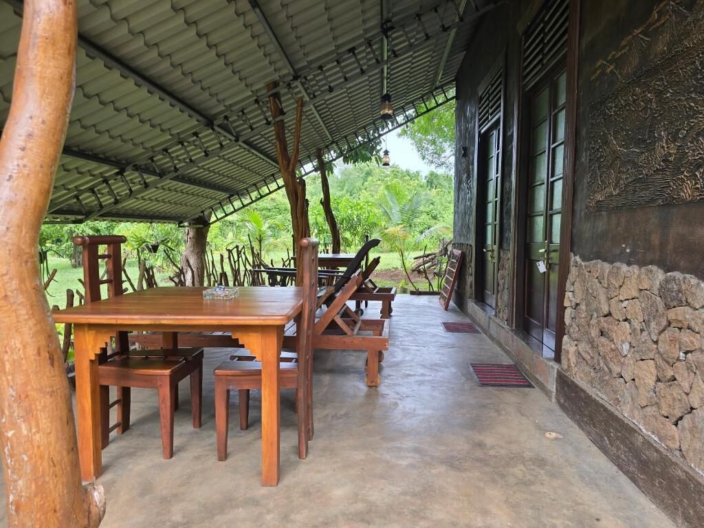 Un área de comedor al aire libre, cubierta, con una mesa y sillas de madera, un muro de piedra y vistas a la vegetación.