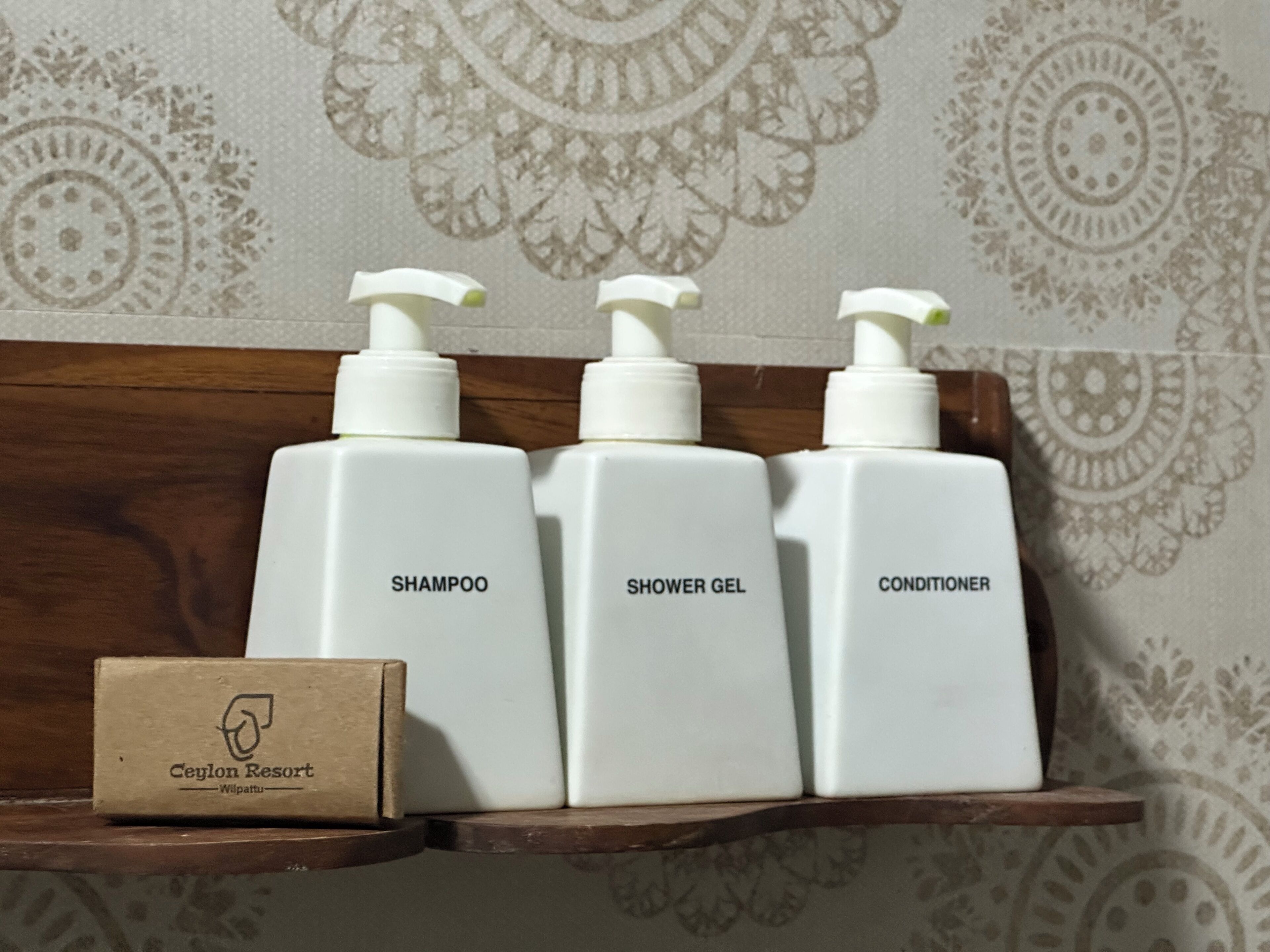 Three white bottles labeled shampoo, shower gel, and conditioner on a wooden shelf.