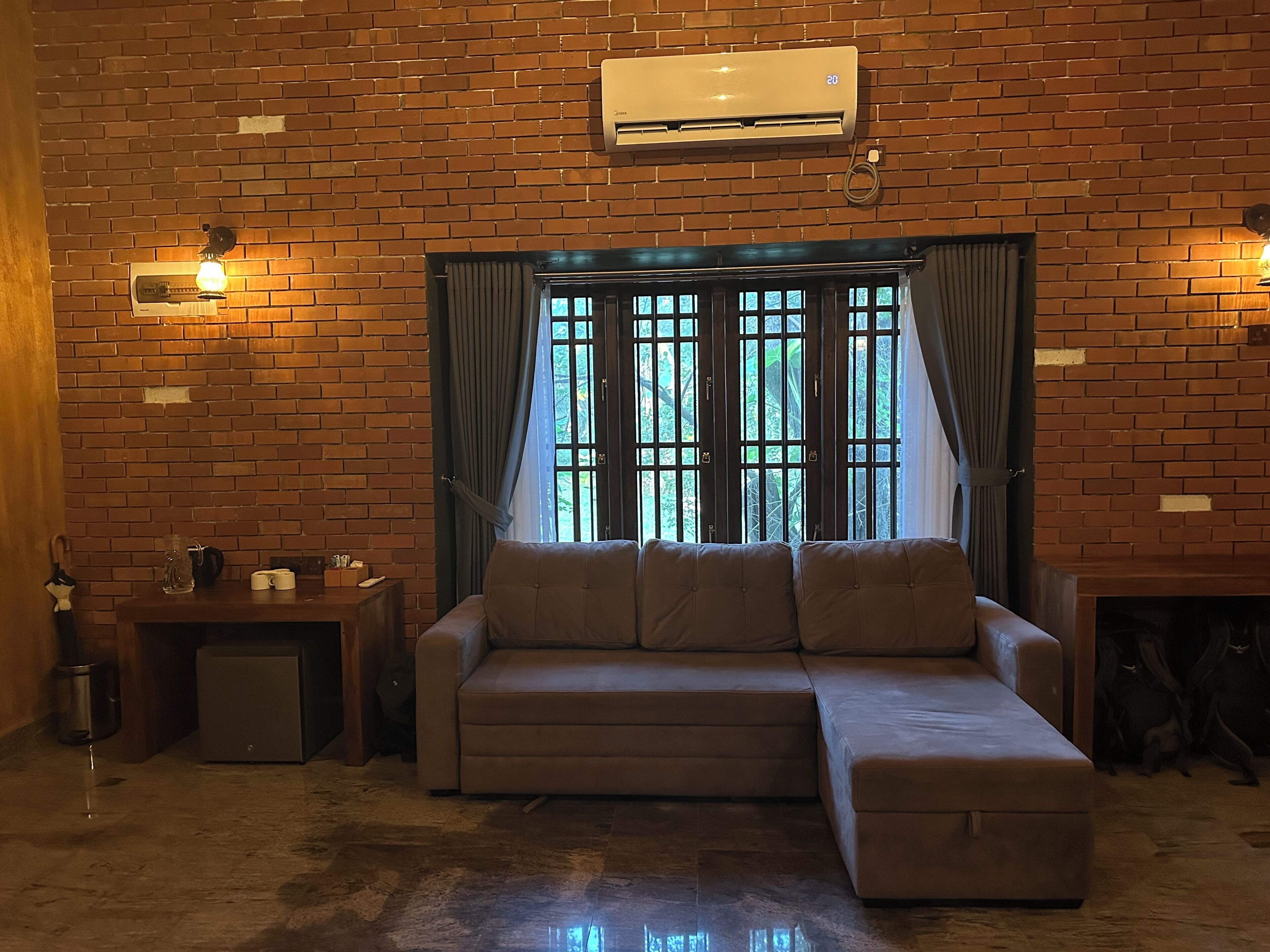 Una habitación con pared de ladrillo, un sofá, una ventana con cortinas, un equipo de aire acondicionado y una pequeña mesa con lámpara.
