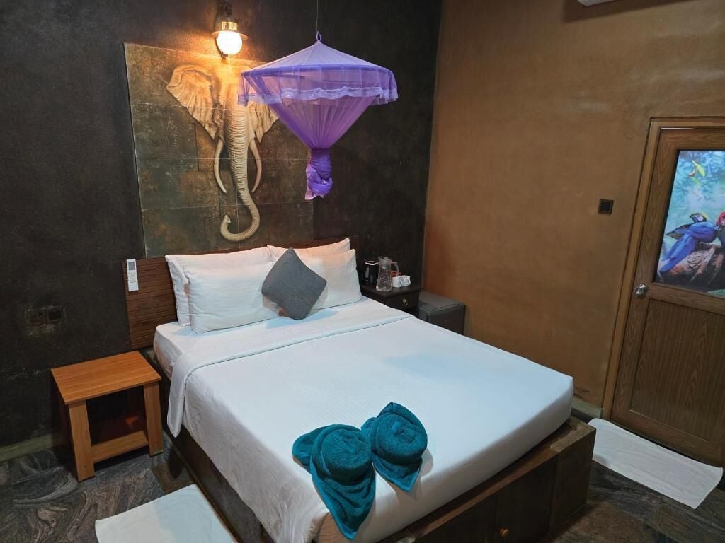 A hotel room with a bed, a nightstand, a wall-mounted elephant head decoration, a purple canopy, and a framed picture on the wall.