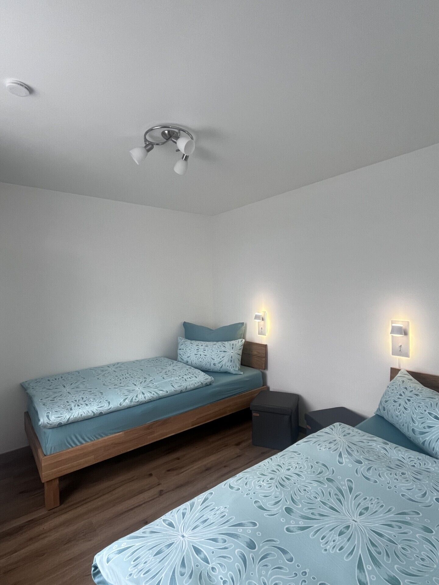 Una habitación con una cama de madera, una mesita de noche pequeña, una lámpara fijada a la pared y un ventilador de techo.