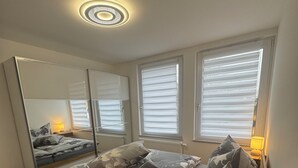 Ein Schlafzimmer mit einem Bett, Nachttischlampen, einem Fenster mit Jalousien und einer Deckenleuchte.