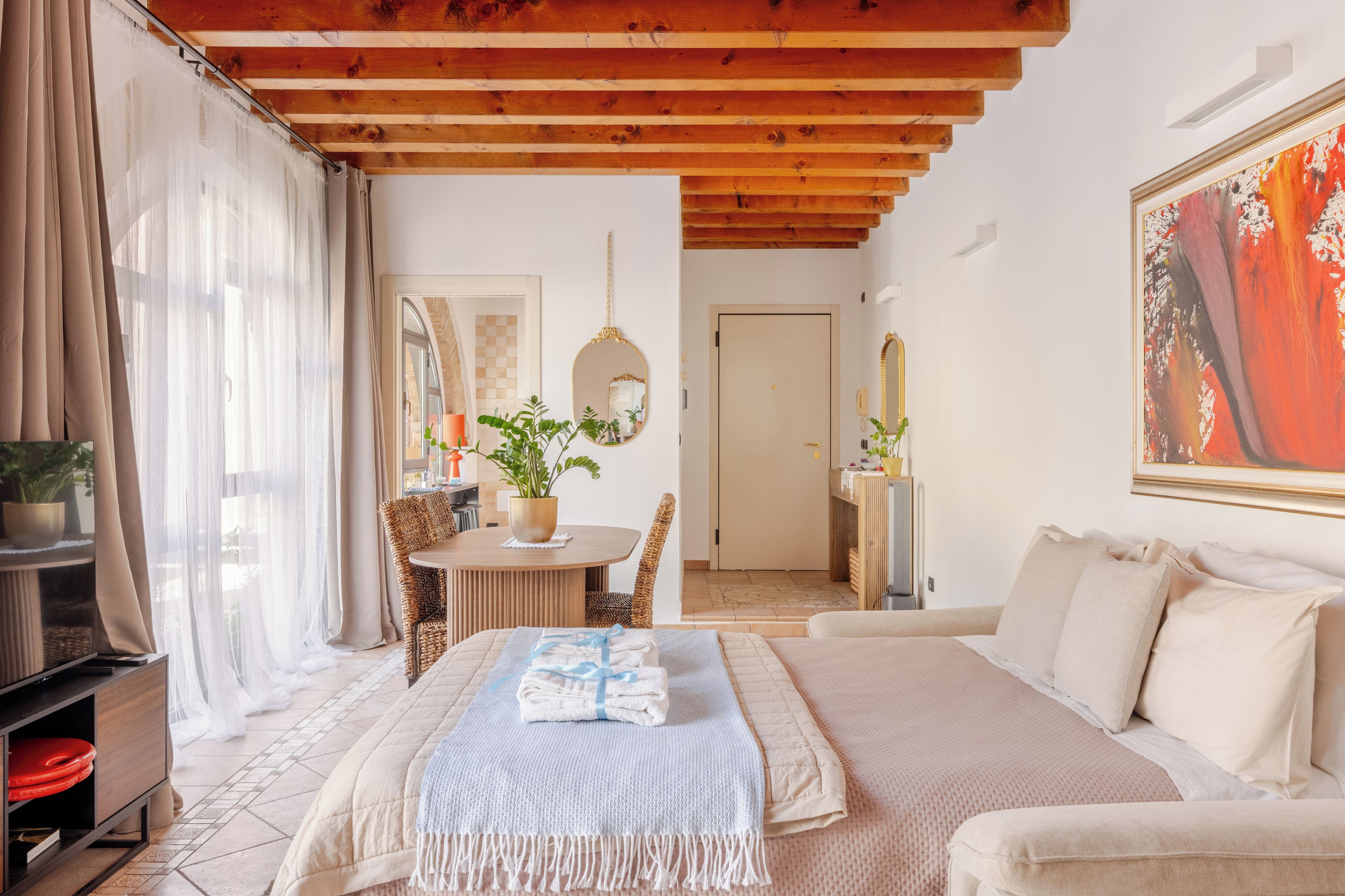 A hotel room with a bed, a sofa, a dining table with chairs, a mirror, and a painting on the wall.