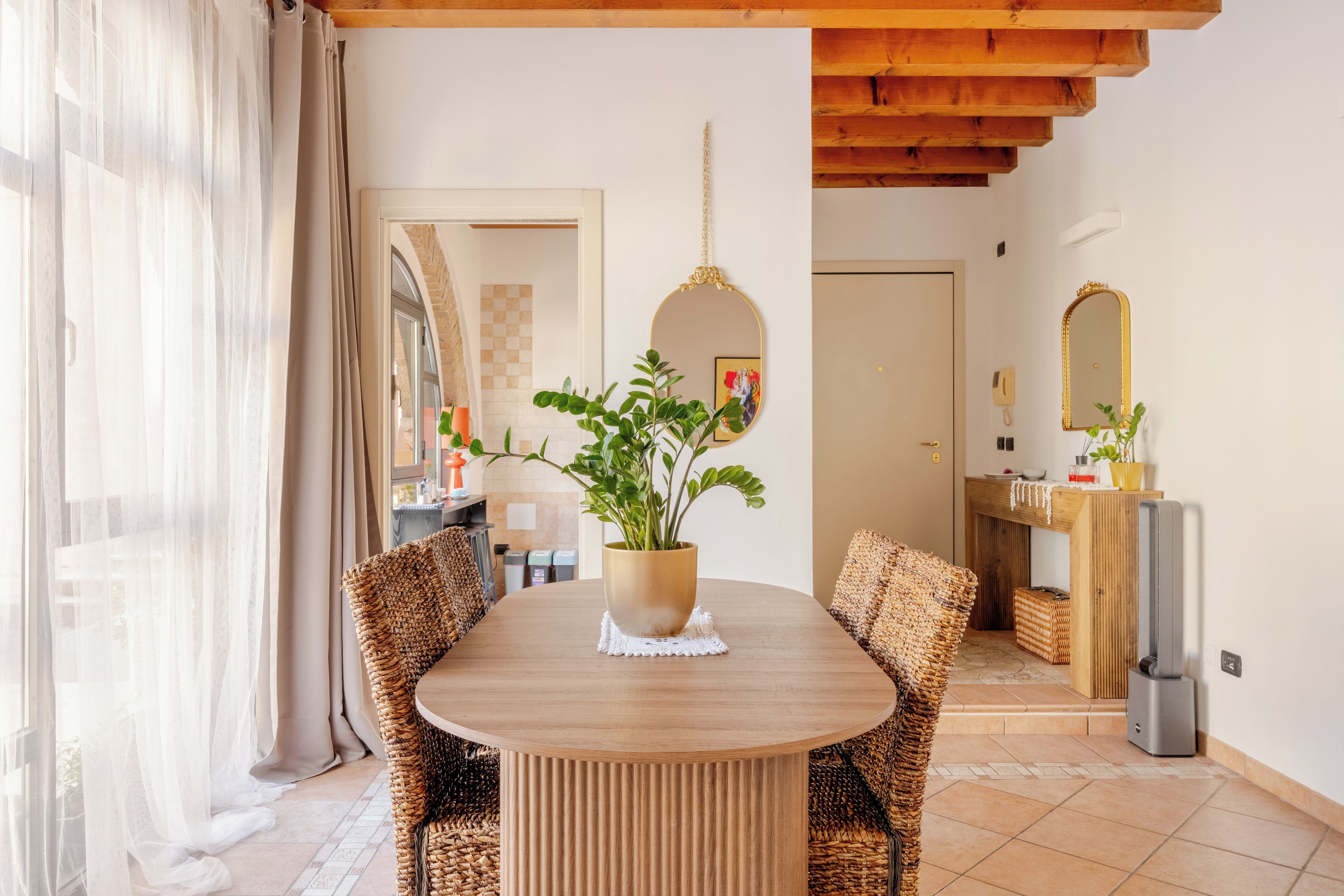 A dining area with a round wooden table, wicker chairs, a potted plant, and a mirror on the wall.