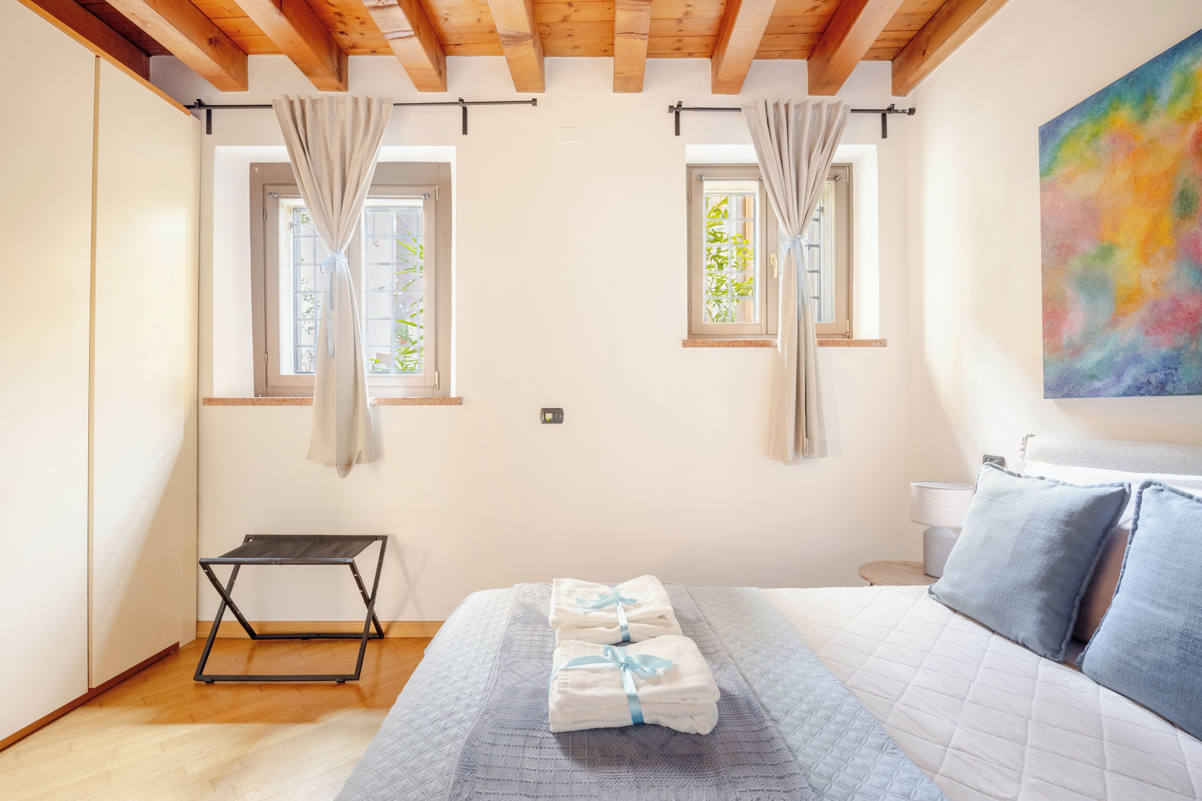 Una camera da letto con un letto, un tavolino, due finestre con tende e un quadro appeso al muro.