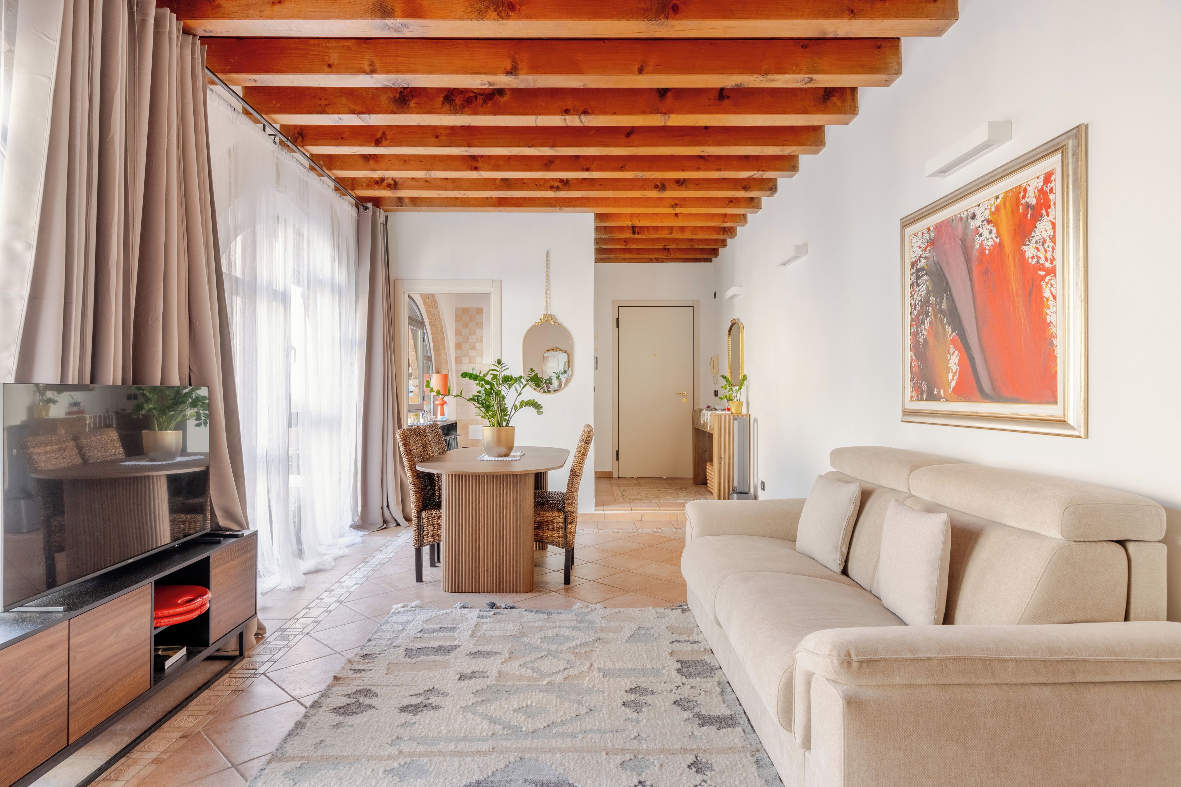 Ein Wohnzimmer mit Holzbalkendecke, einem beigen Sofa, einem Esstisch mit Stühlen, einem Fernseher auf einem Holzboden und einem großen Gemälde an der Wand.