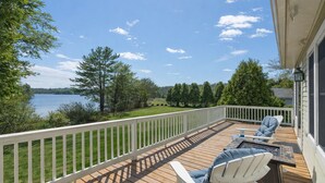 Um terraço com duas cadeiras Adirondack e uma mesa, com vista para um lago rodeado por árvores e uma área gramada.