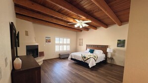 Quarto com teto de madeira, cama com cabeceira, mesa de cabeceira, luminária e janela com persianas.