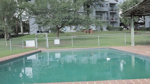 A swimming pool with a brick border, surrounded by a white fence, trees, and a building with balconies in the background. - Luxurious, Spacious 2-br (Johannesburg)