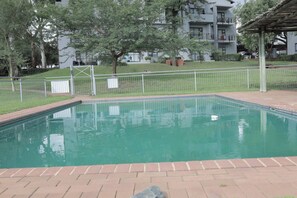 A swimming pool with a brick border, surrounded by a white fence, trees, and a building with balconies in the background. - Luxurious, Spacious 2-br (Johannesburg)