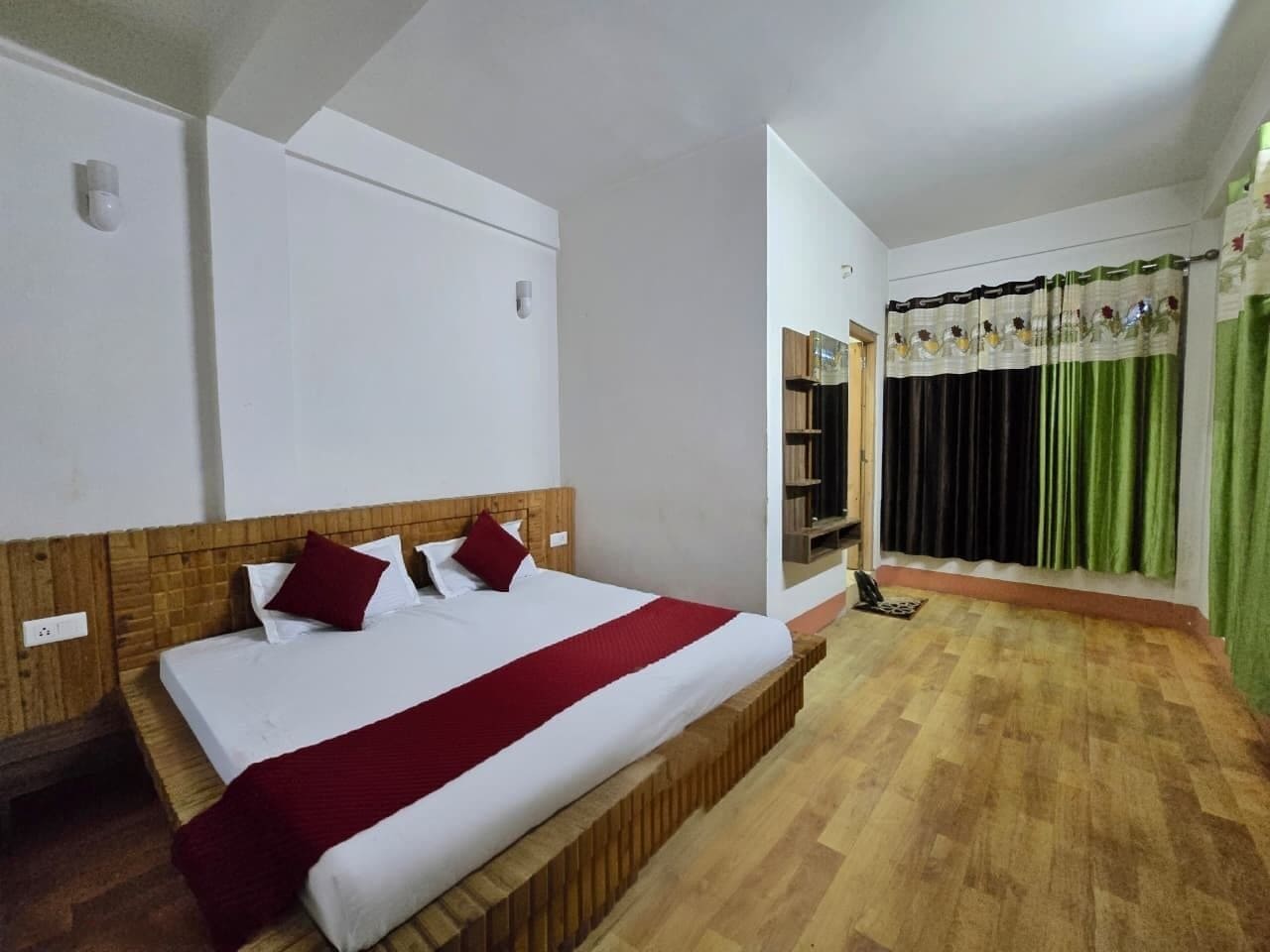 A hotel room with a wooden bed, white walls, a wooden headboard, a red bedspread, and a green curtain.