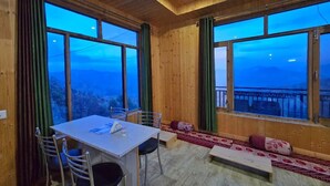 A wooden interior with a table and chairs, large windows with curtains, and a view of a mountainous landscape. - Lucky Homestay (Theog)