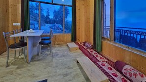 A wooden interior with a dining table and chairs, a bench with cushions, and large windows offering a view of the outdoors. - Lucky Homestay (Theog)