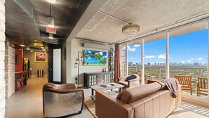 Ein modernes Wohnzimmer mit Sofa, Sessel und Fernseher. Ein Balkon bietet Blick auf die Stadt.