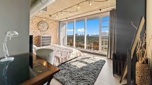 Ein modernes Schlafzimmer mit einem großen Bett, einem Schreibtisch, einem Stuhl, einem Fenster mit Blick auf die Stadt und einer Ziegelwand.