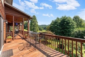 Una terraza de madera con mobiliario exterior, una parrilla y una vista panorámica de un valle boscoso.