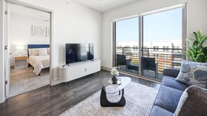 A modern living room with a large window, a ceiling fan, a flat-screen TV, and a sofa. - Modern Oasis + Pool, Gym + EV Charger (Frisco)