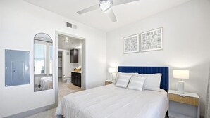A bedroom with a bed, a ceiling fan, a nightstand, a mirror, and a door leading to another room. - Modern Oasis + Pool, Gym + EV Charger (Frisco)
