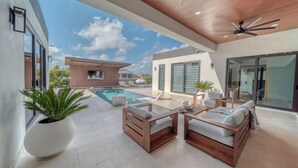 A modern outdoor living area with a pool, wooden furniture, and a ceiling fan. - Cozy Modern Villa - Heated Pool & Spa - BBQ - Water View (Cape Coral)