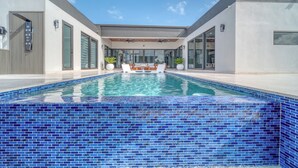A modern pool area with a clear blue mosaic tile border, a seating area with white furniture, and a contemporary building with large windows and a flat roof. - Cozy Modern Villa - Heated Pool & Spa - BBQ - Water View (Cape Coral)