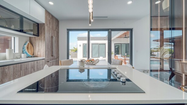 A modern kitchen with a central island, wooden cabinetry, and a view to the outdoor seating area. - Cozy Modern Villa - Heated Pool & Spa - BBQ - Water View (Cape Coral)