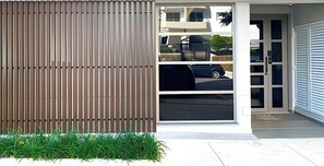 Entrada moderna de un edificio con puerta corredera de vidrio, revestimiento de listones de madera verticales y un pequeño espacio con plantas verdes.