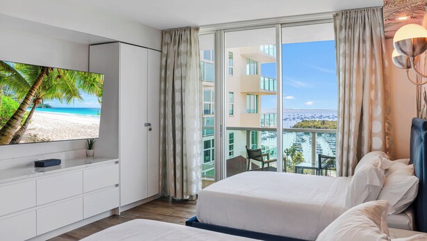 Et hotelværelse med et fjernsyn, der viser et strandbillede, en balkon med havudsigt og bygninger, samt en seng med hvide sengetæpper.