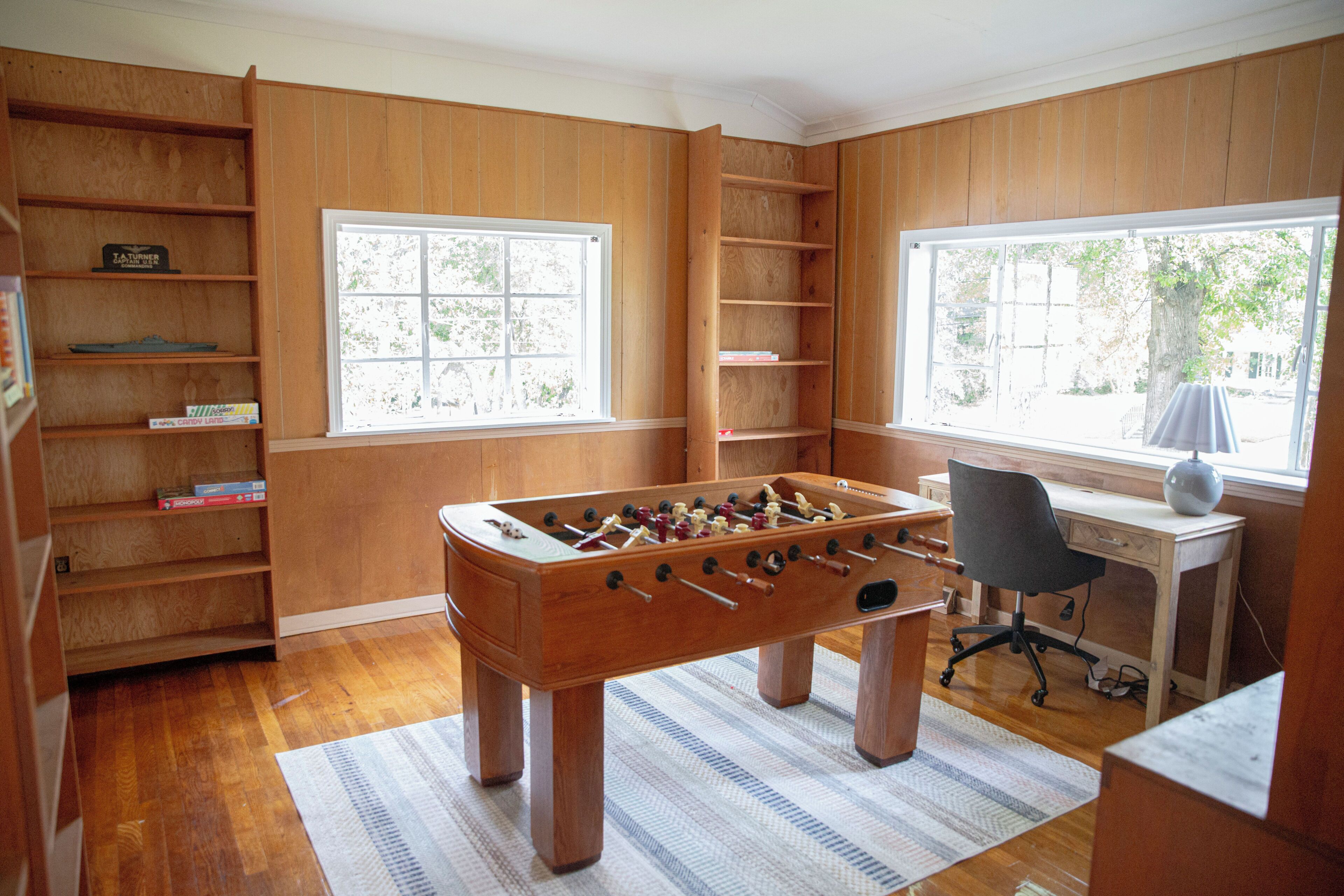 Una habitación con paneles de madera, una mesa de fútbol de mesa, un escritorio con una silla y ventanas grandes.