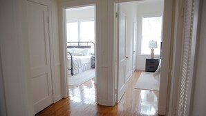 Corredor que dá para um quarto com cama, mesa de cabeceira e abajur.
