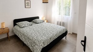 Une chambre à coucher comprenant un lit, une table de chevet, un luminaire suspendu, une fenêtre avec des rideaux et un radiateur fixé au mur.