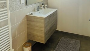 Une salle de bain avec un lavabo blanc, un meuble-lavabo en bois, un miroir, un porte-serviettes, une petite fenêtre et un sol foncé.