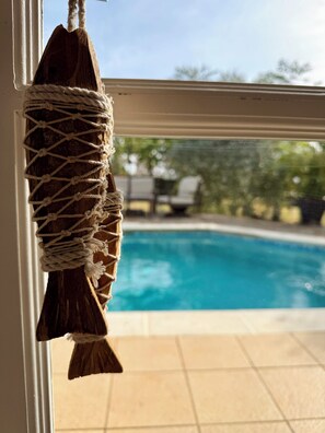 Une décoration en macramé représentant un poisson, suspendue près d’une fenêtre donnant sur une piscine et sur du mobilier d’extérieur.