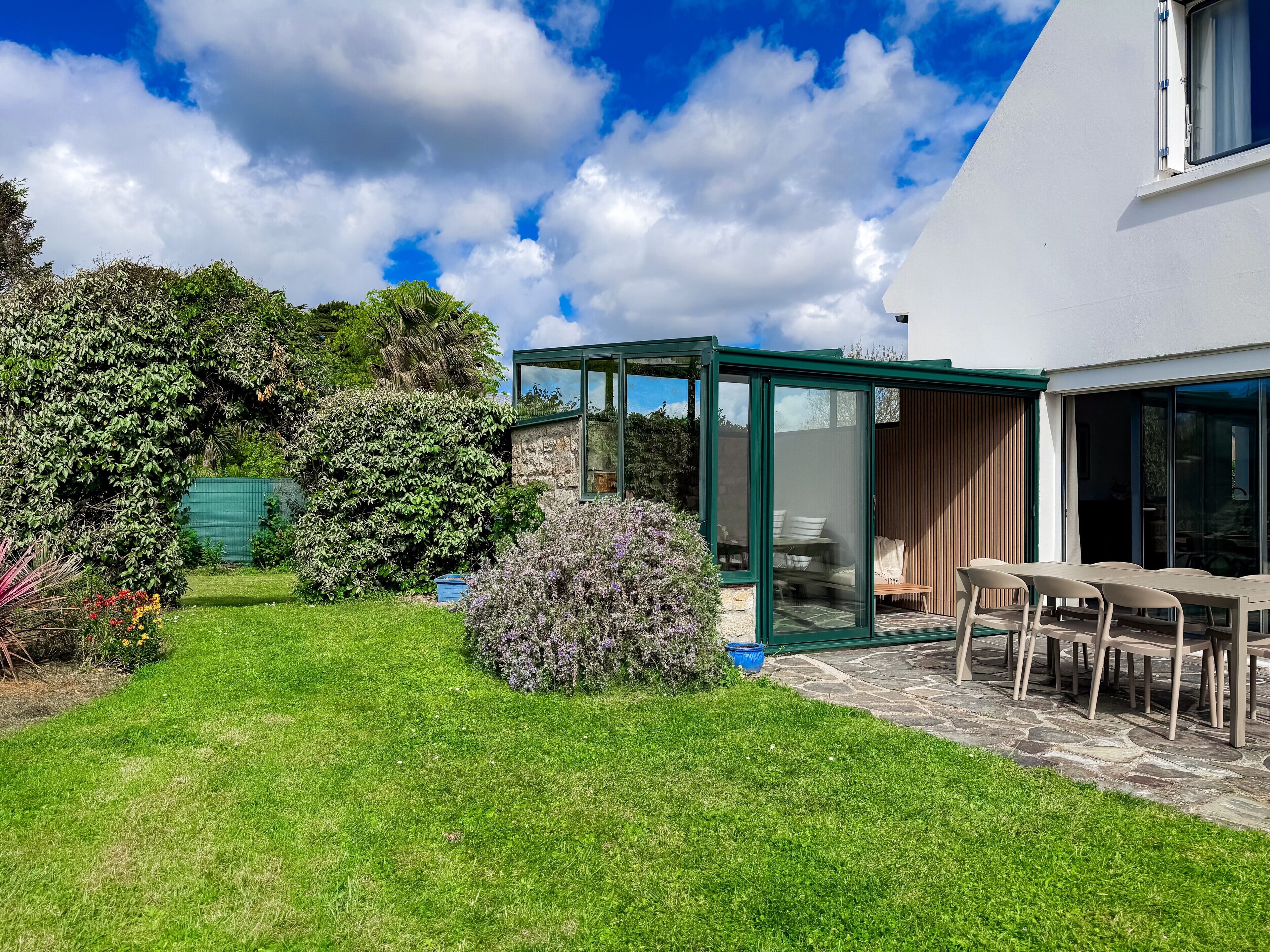 A modern house with a glass conservatory, a patio with a table and chairs, and a well-maintained garden.