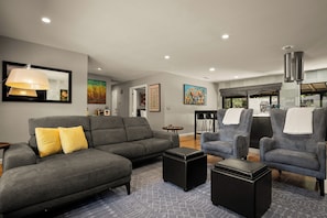 Ein modernes Wohnzimmer mit grauem Sofa, Hocker und Sesseln, einem Essbereich mit Stühlen und einer Küche im Hintergrund.