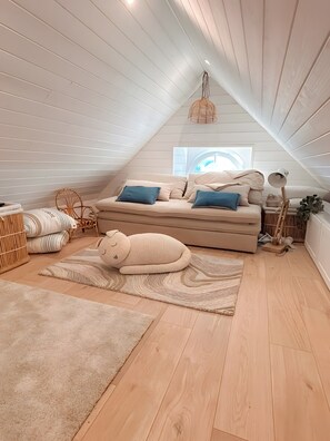 A cozy attic bedroom with a sloped ceiling, a large window, a sofa with cushions, a decorative lamp, and a plush toy.