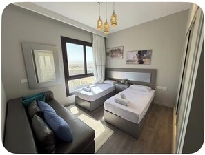 A modern hotel room with a sofa, two beds, a large window, and a city view. - El-Gouna Gem
 Tranquil 3-bedroom apartment with AC in Red Sea Governorate (Hurghade 2)