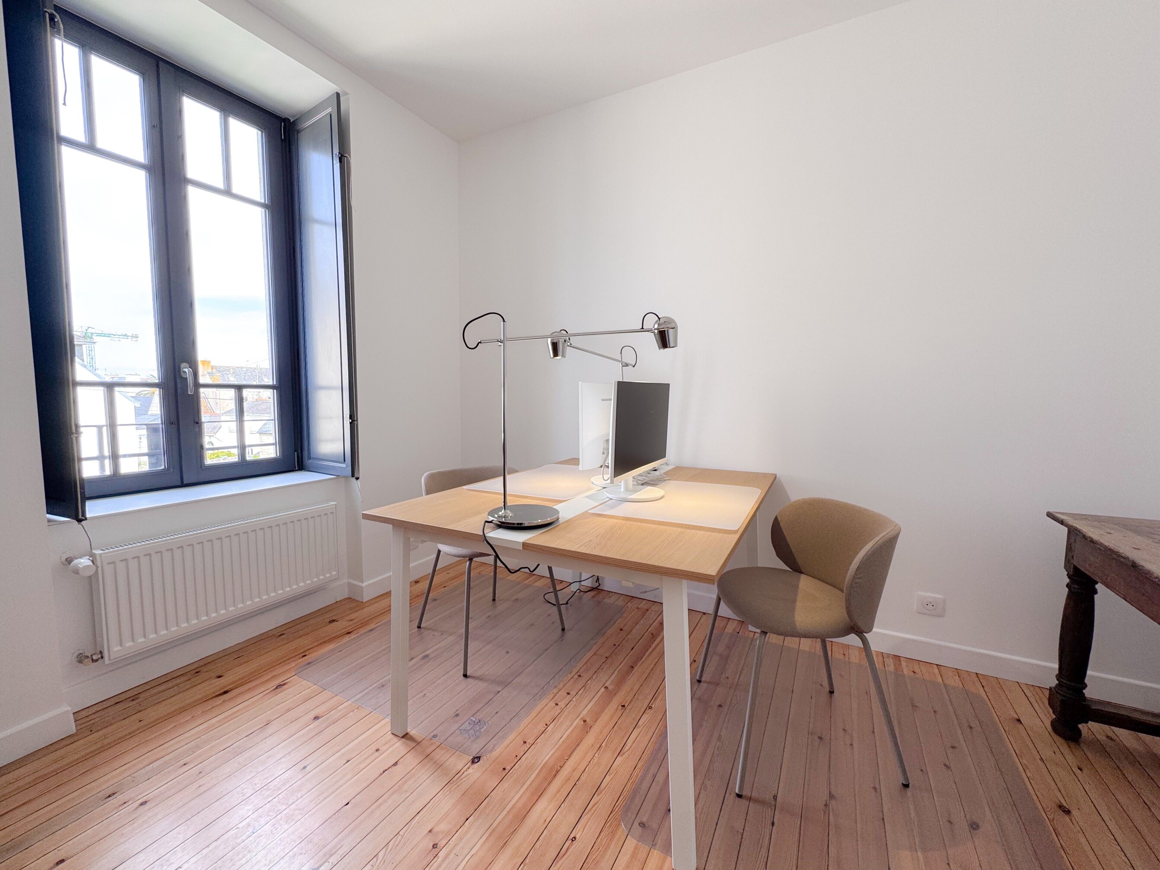 A room with a wooden floor, a desk with a computer and a lamp, a chair, and a window with blinds.