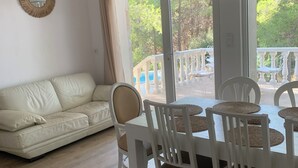 Ein modernes Wohnzimmer mit einem weißen Sofa, einem Esstisch mit weißen Stühlen, Weidenpendelleuchten und Blick ins Grüne.